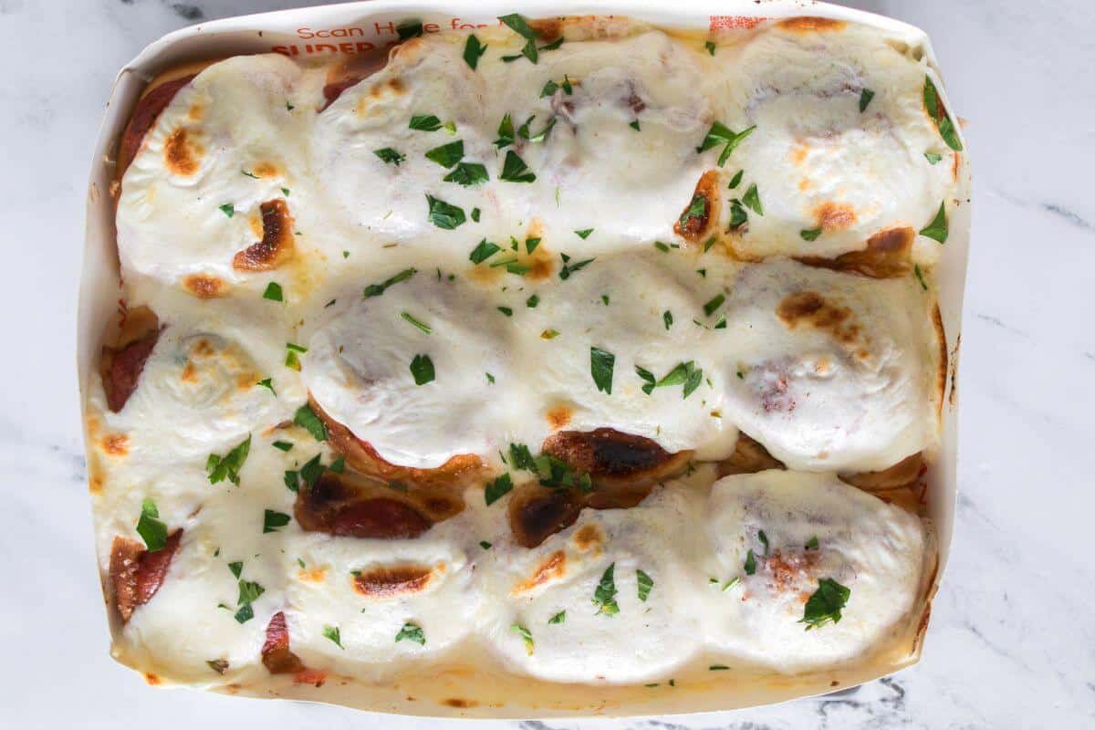 A tray of baked manicotti covered in melted cheese and garnished with chopped parsley, photographed from above on a marble surface.