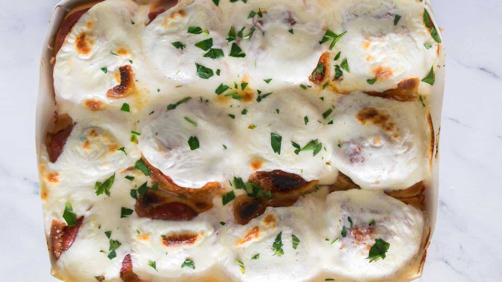 A baking dish filled with meatballs covered in melted cheese and garnished with chopped parsley on a white surface.