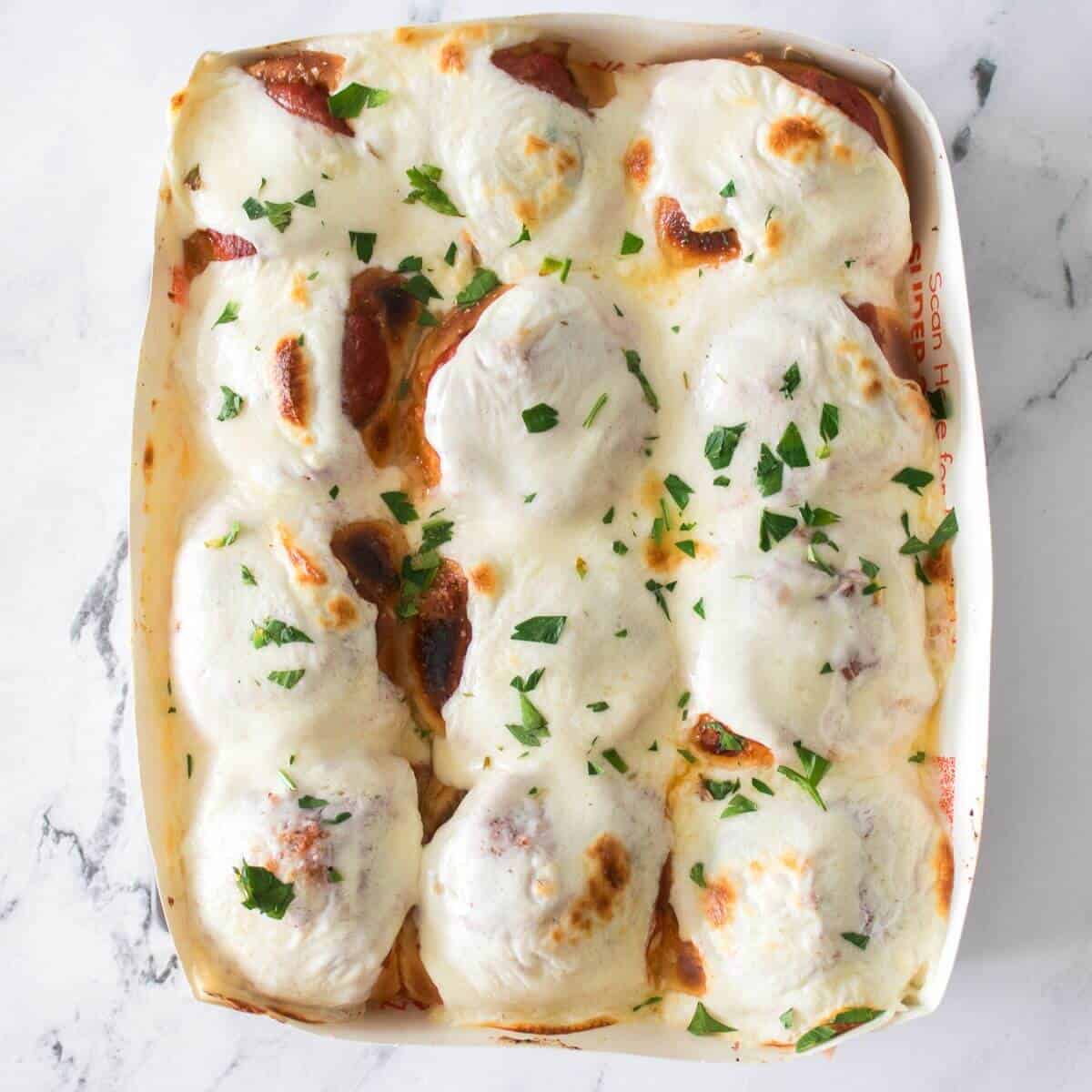 A tray of baked meatballs topped with melted mozzarella cheese and garnished with chopped parsley on a marble surface.