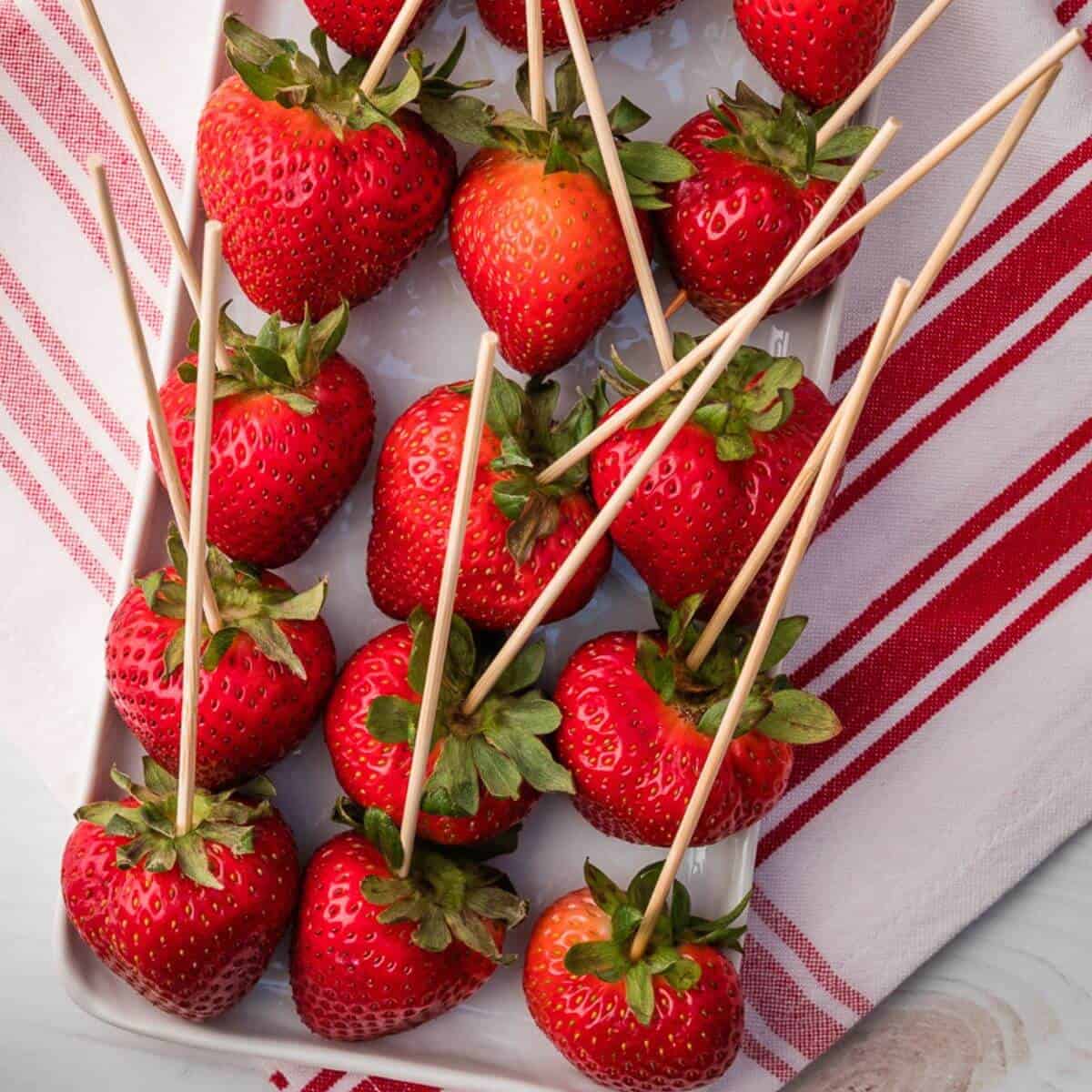 Fresh strawberries on skewers are arranged on a white rectangular plate, set on a white cloth with red stripes.