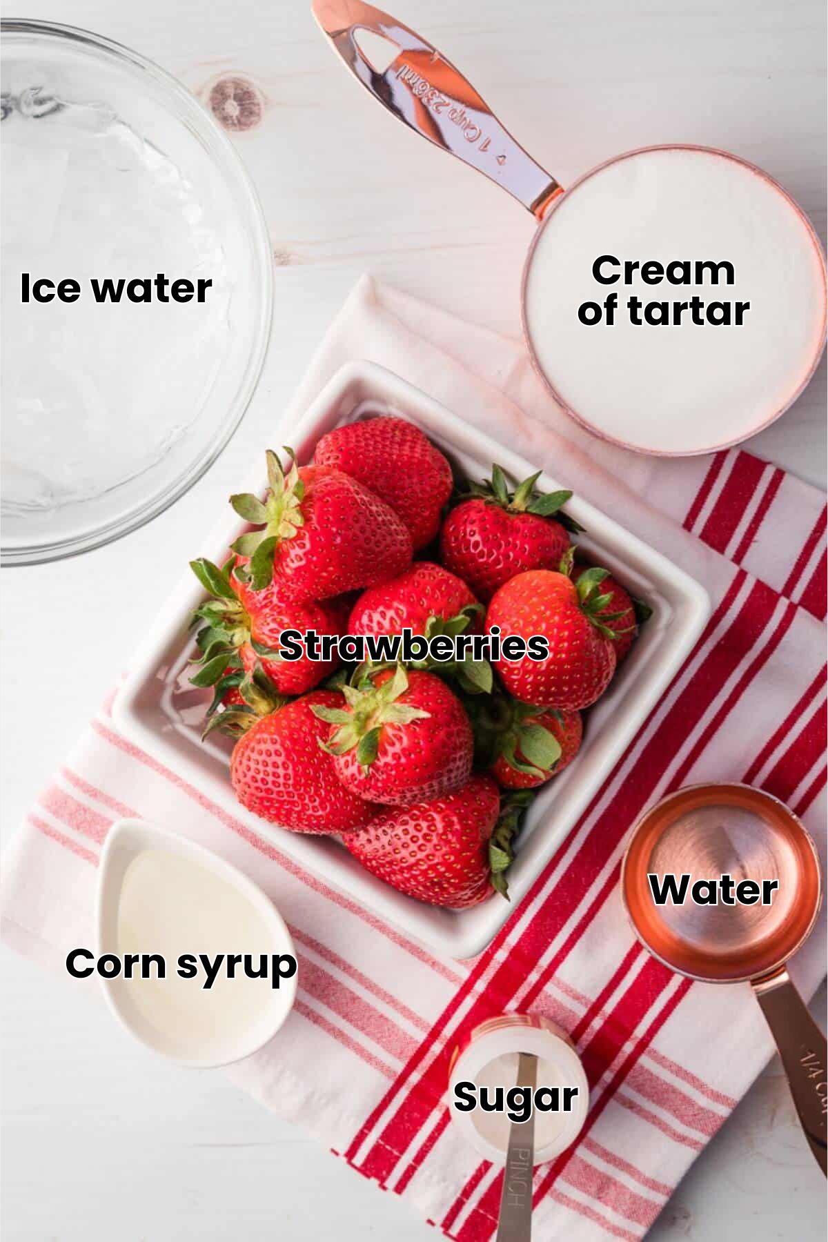 A square bowl of fresh strawberries, a bowl of ice, a cup of sugar, a small pitcher of cream, and measuring spoons on a red-striped towel.