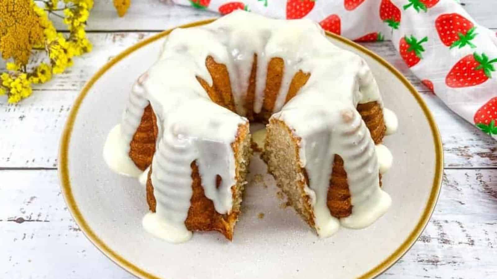A Bundt cake topped with white icing sits on a plate, with one large slice cut out; a floral and strawberry-patterned cloth is nearby.