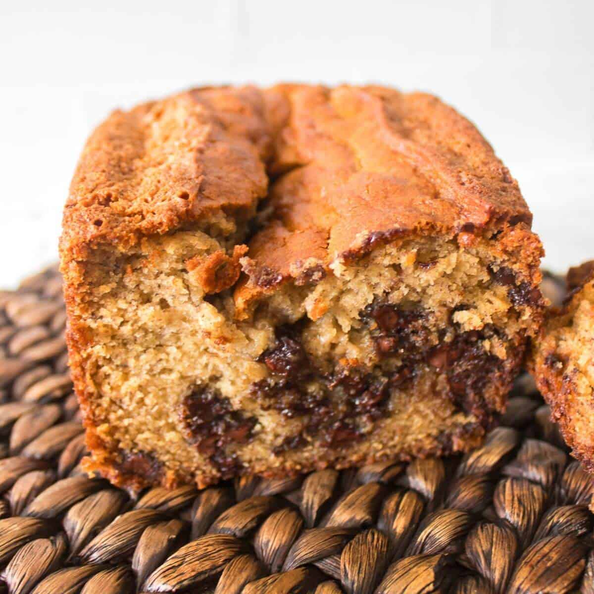 A close-up of a sliced loaf of banana bread with visible chocolate chips on a woven placemat.