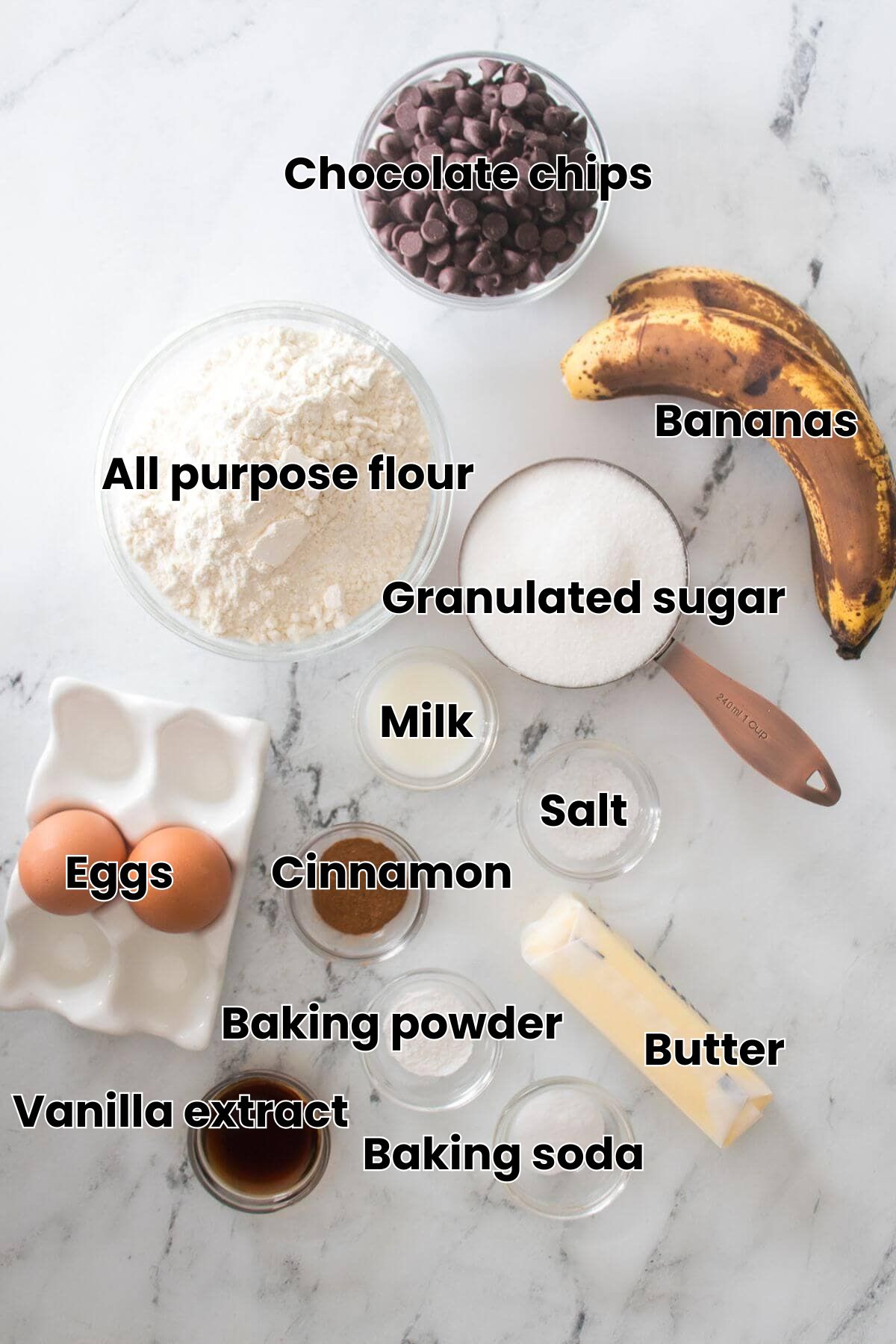 Baking ingredients on a marble surface, including flour, sugar, chocolate chips, overripe bananas, eggs, butter, and small bowls of milk, cinnamon, vanilla, baking powder, and salt.