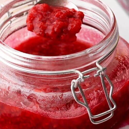 An open glass jar filled with raspberry sauce sits on a pink checkered cloth, with a spoon lifting some sauce out of the jar.