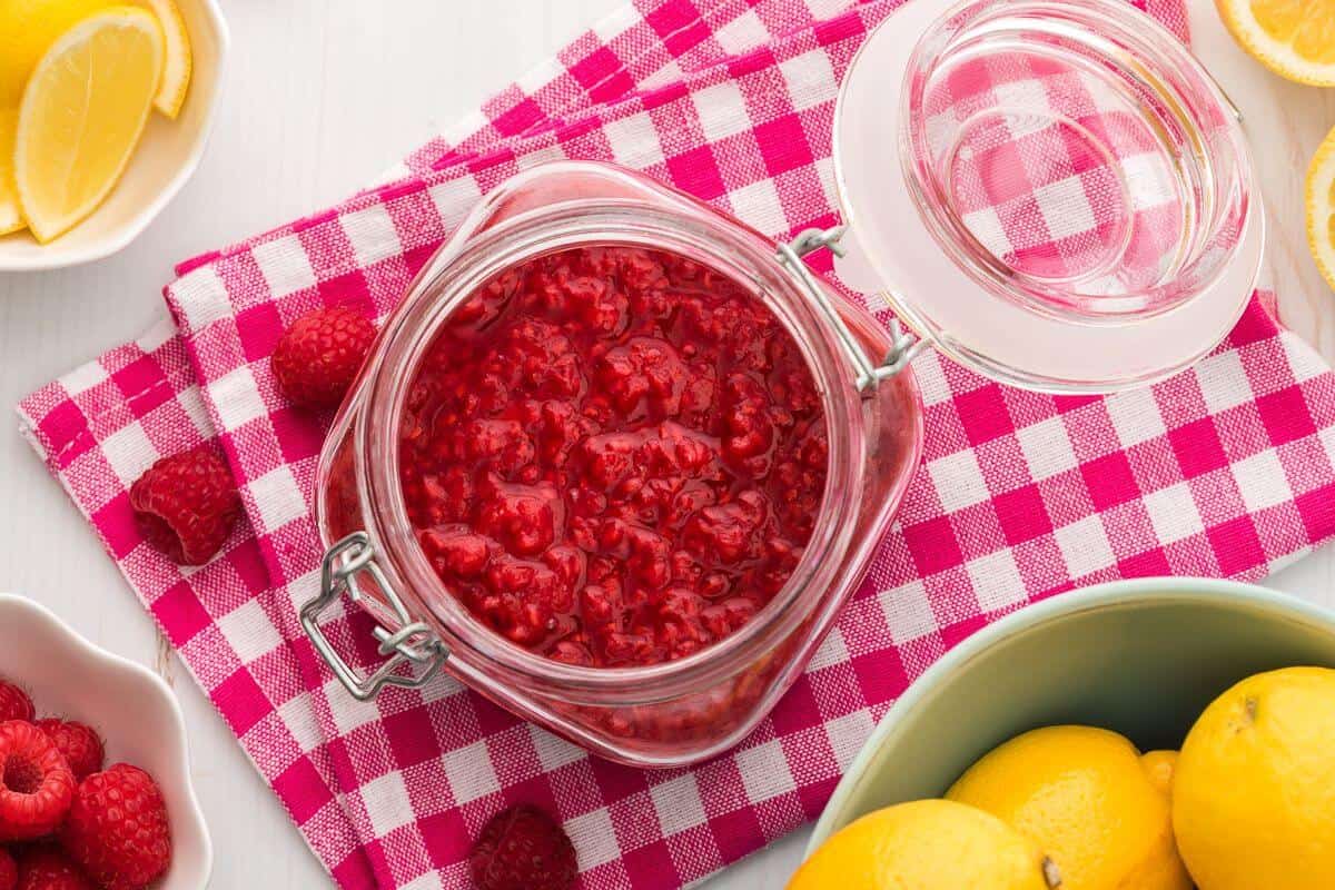 An open jar of raspberry jam sits on a red and white checkered cloth, surrounded by fresh raspberries and whole lemons.