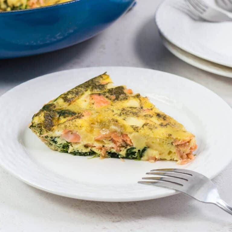 A slice of frittata with spinach and salmon on a white plate, with a fork placed in front and extra plates in the background.