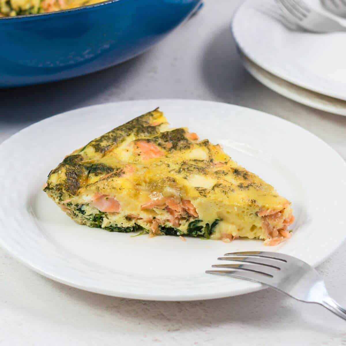 A slice of frittata with spinach and salmon on a white plate, with a fork placed in front and extra plates in the background.