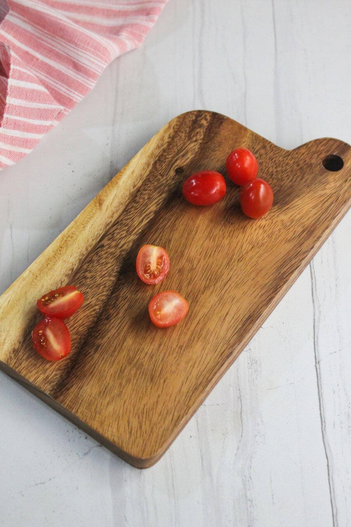 A wooden cutting board with whole and halved grape tomatoes on top, placed on a white surface next to a pink striped cloth.