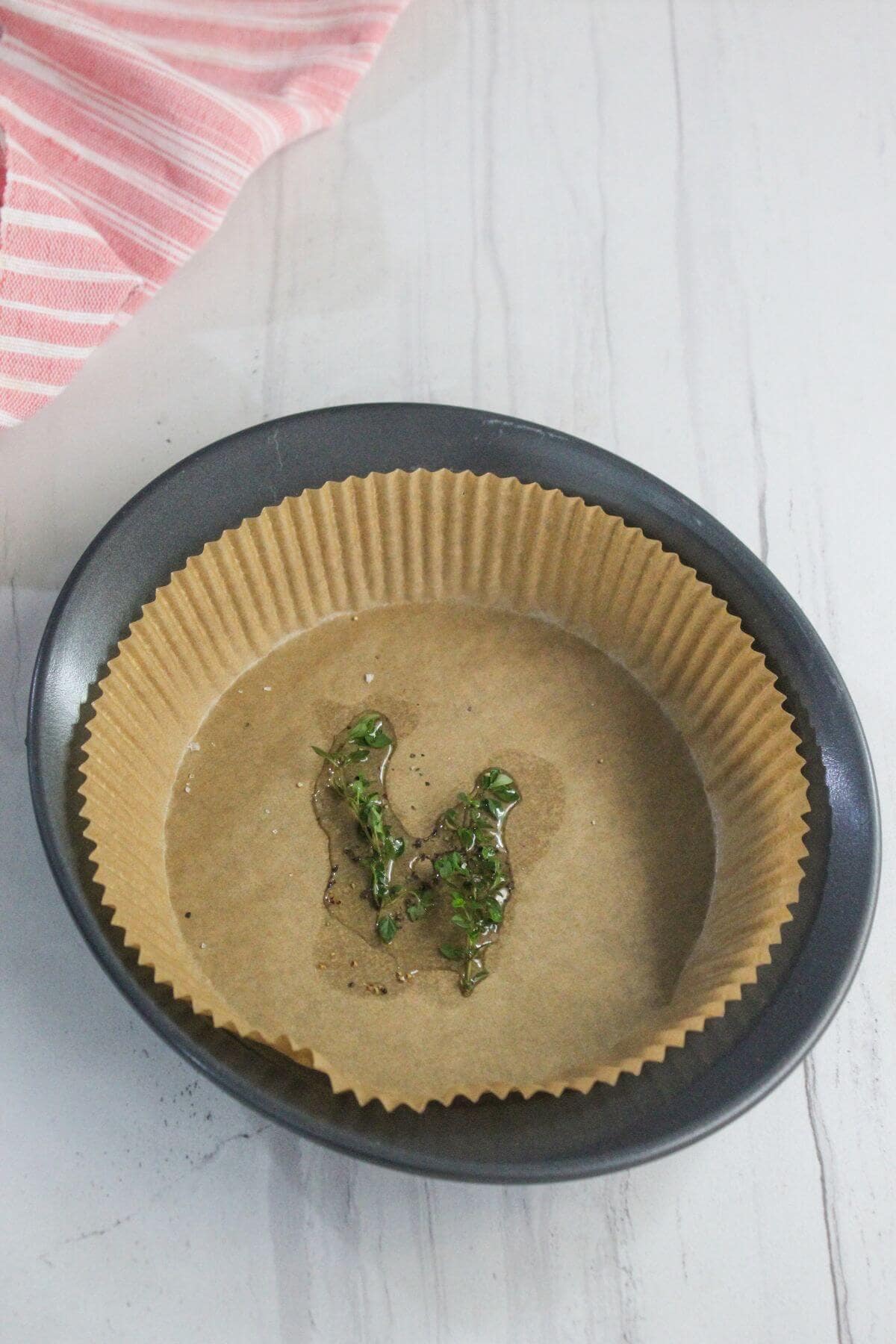 A round baking pan lined with parchment paper holds a small amount of oil and fresh chopped herbs in the center. A pink and white striped cloth is partially visible in the corner.