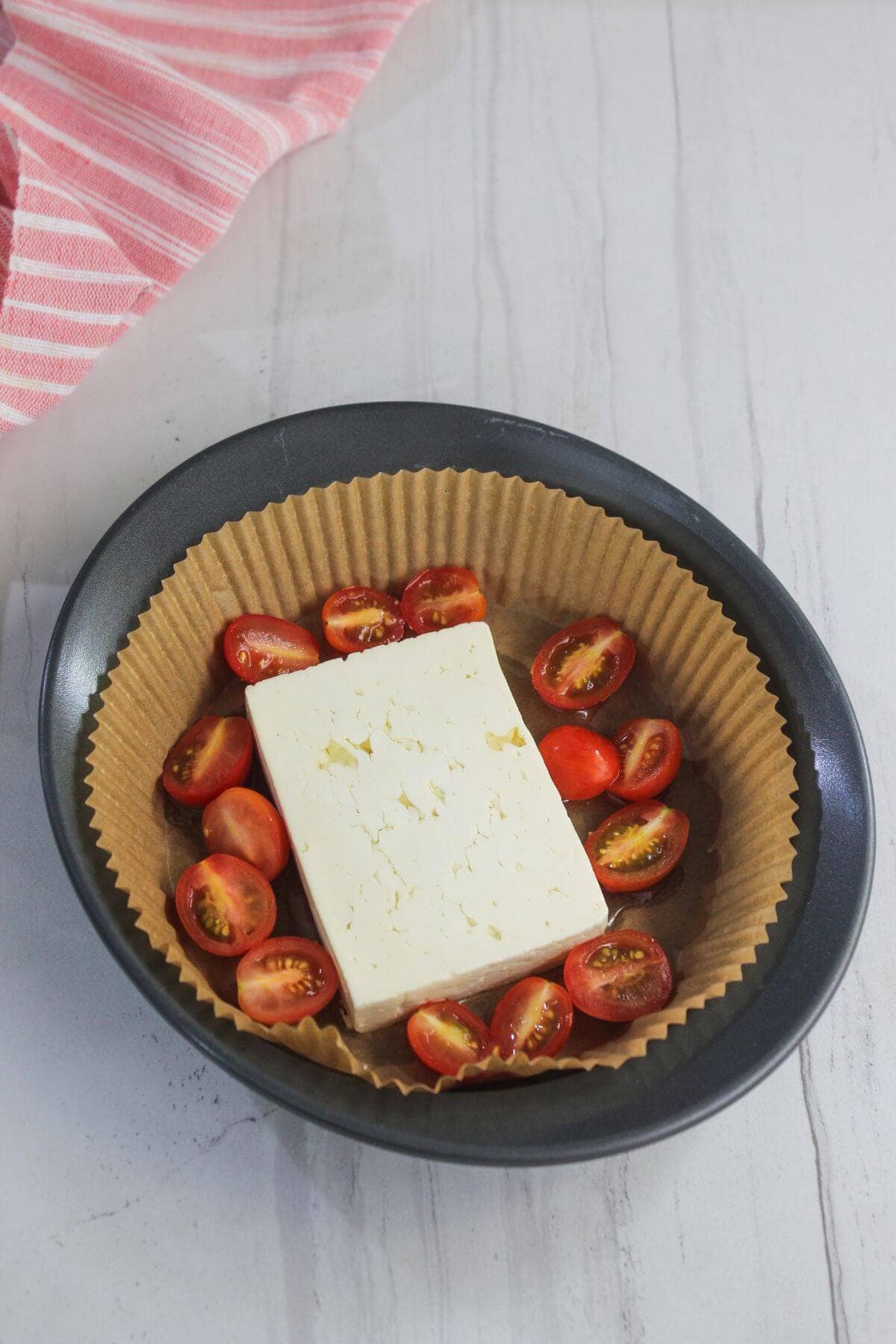 Round baking dish lined with parchment paper, containing a block of feta cheese surrounded by halved cherry tomatoes. A pink striped cloth is partially visible in the corner.