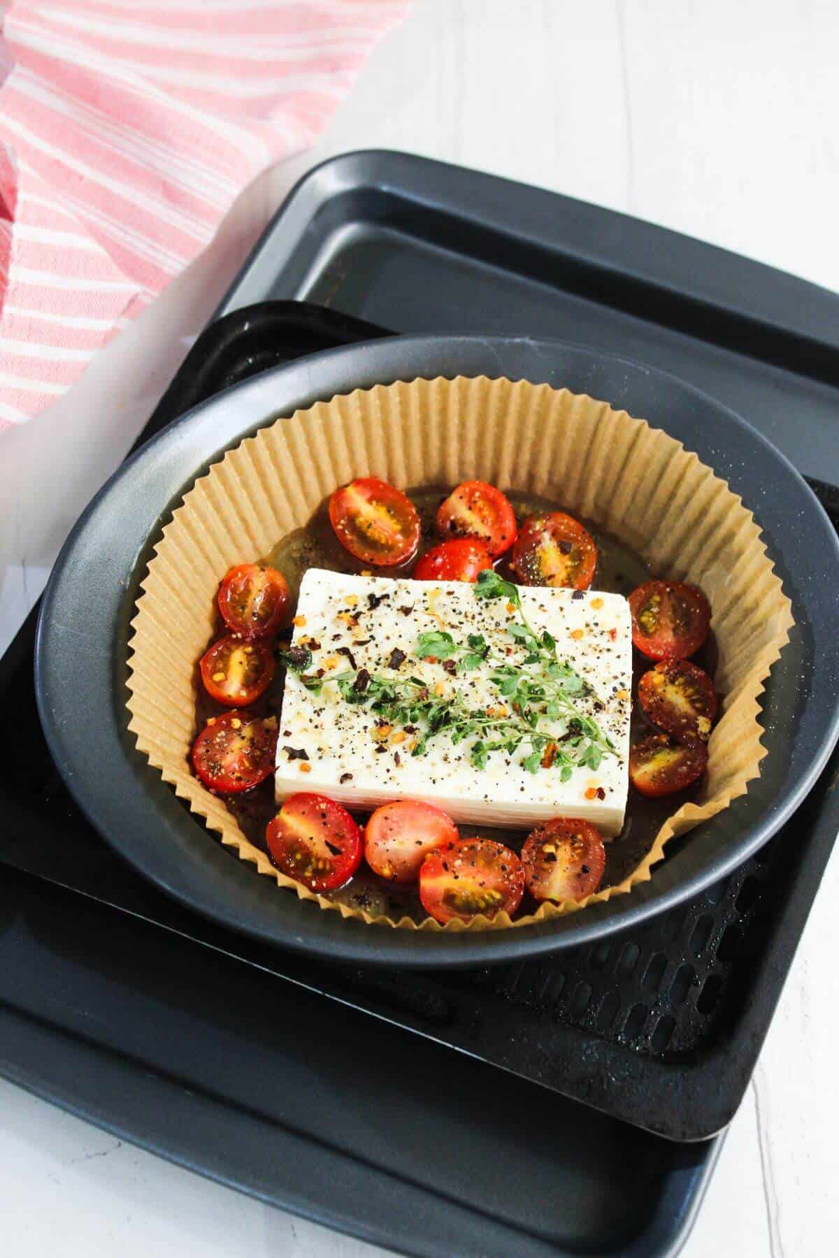 A round baking pan lined with parchment paper contains cherry tomato halves and a block of feta cheese, topped with herbs and pepper, ready to be baked.