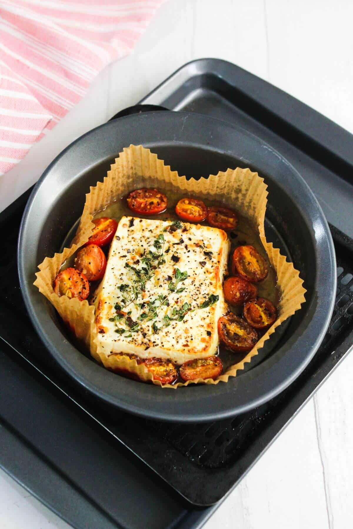 A round baking pan lined with parchment holds a baked feta cheese block topped with herbs, surrounded by roasted cherry tomatoes.