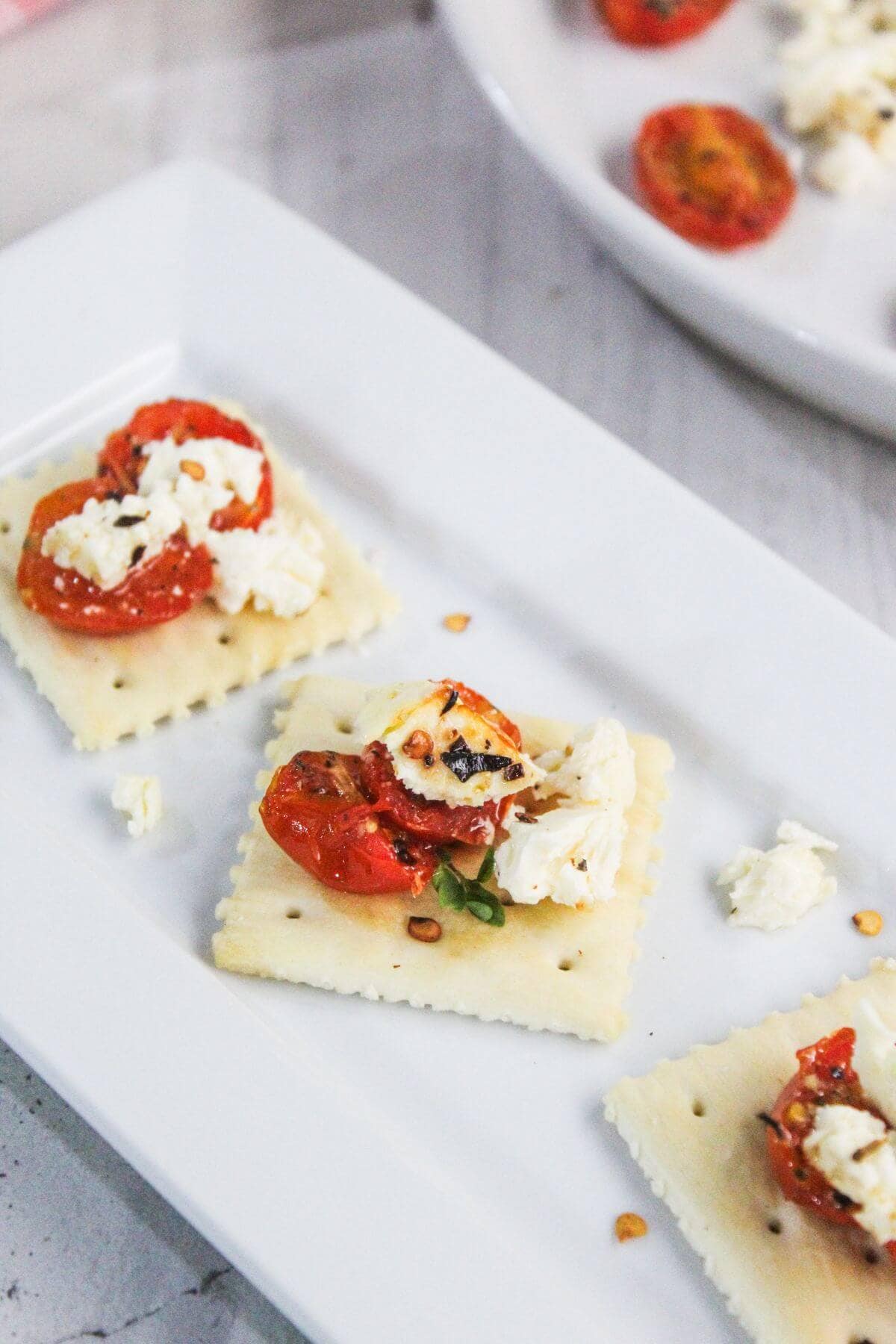 Square crackers topped with roasted tomatoes and crumbled cheese are arranged on a white rectangular plate.