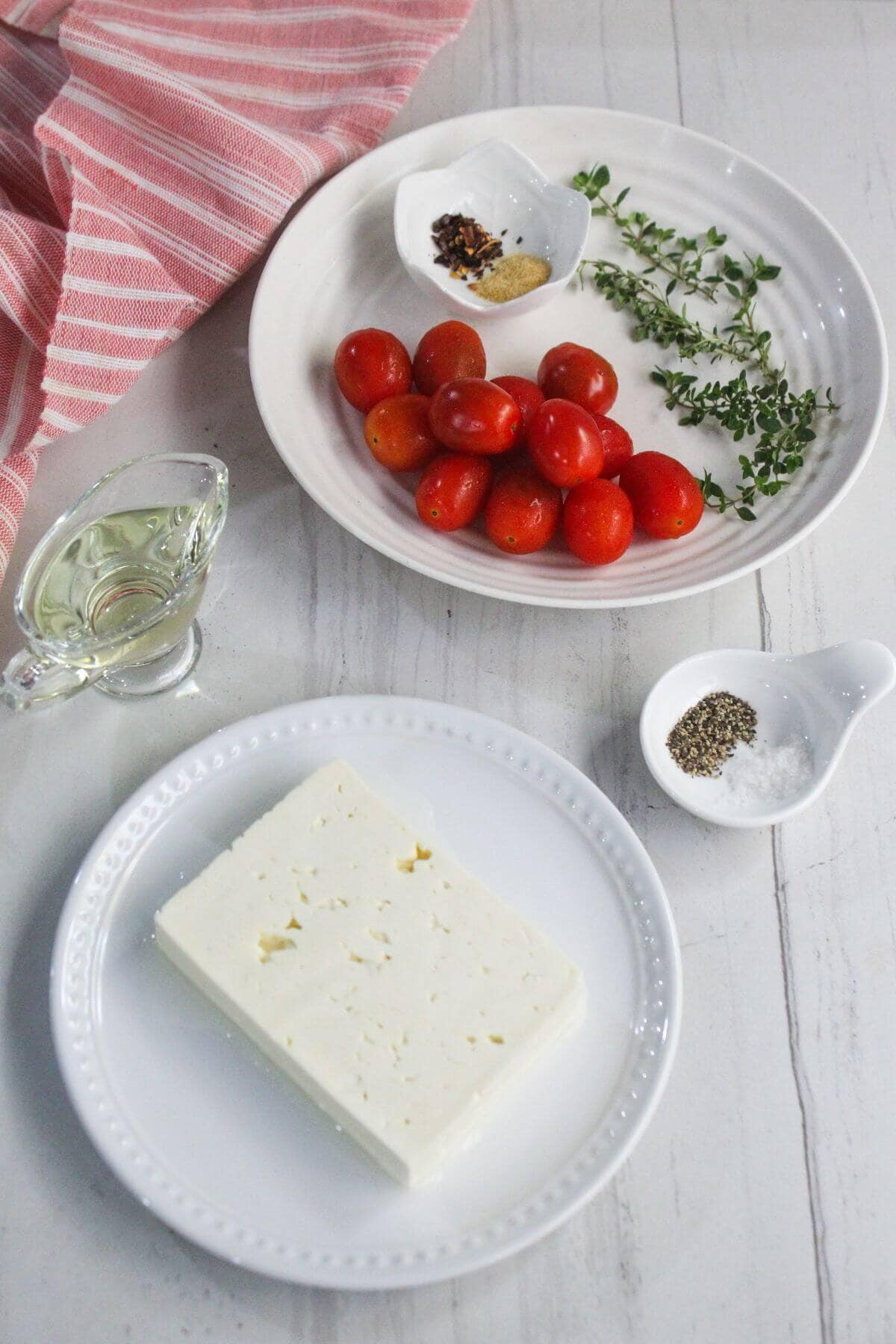A plate of cherry tomatoes, fresh herbs, and spices, a block of feta cheese on a separate plate, a small glass of olive oil, salt, and pepper on a white surface.