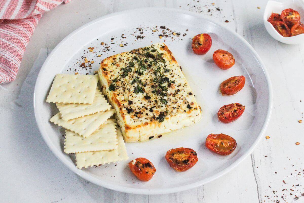 A plate with a baked block of seasoned feta cheese, cherry tomato halves, and square crackers, on a white surface.
