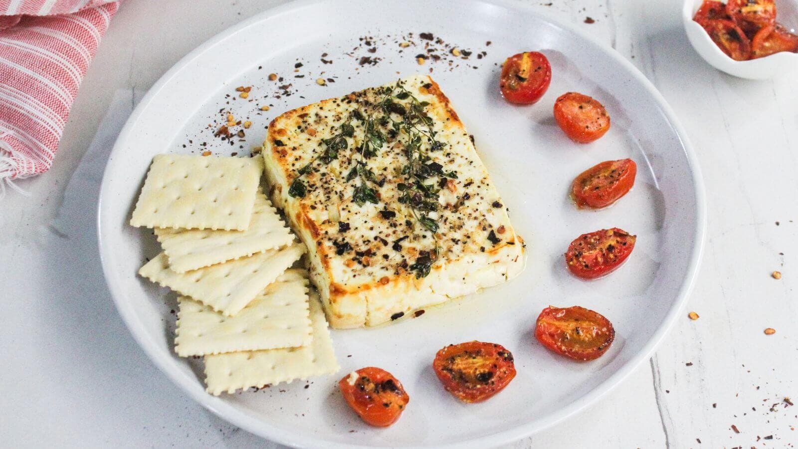 A plate with baked feta cheese topped with herbs and spices, surrounded by cherry tomato halves and several square crackers.