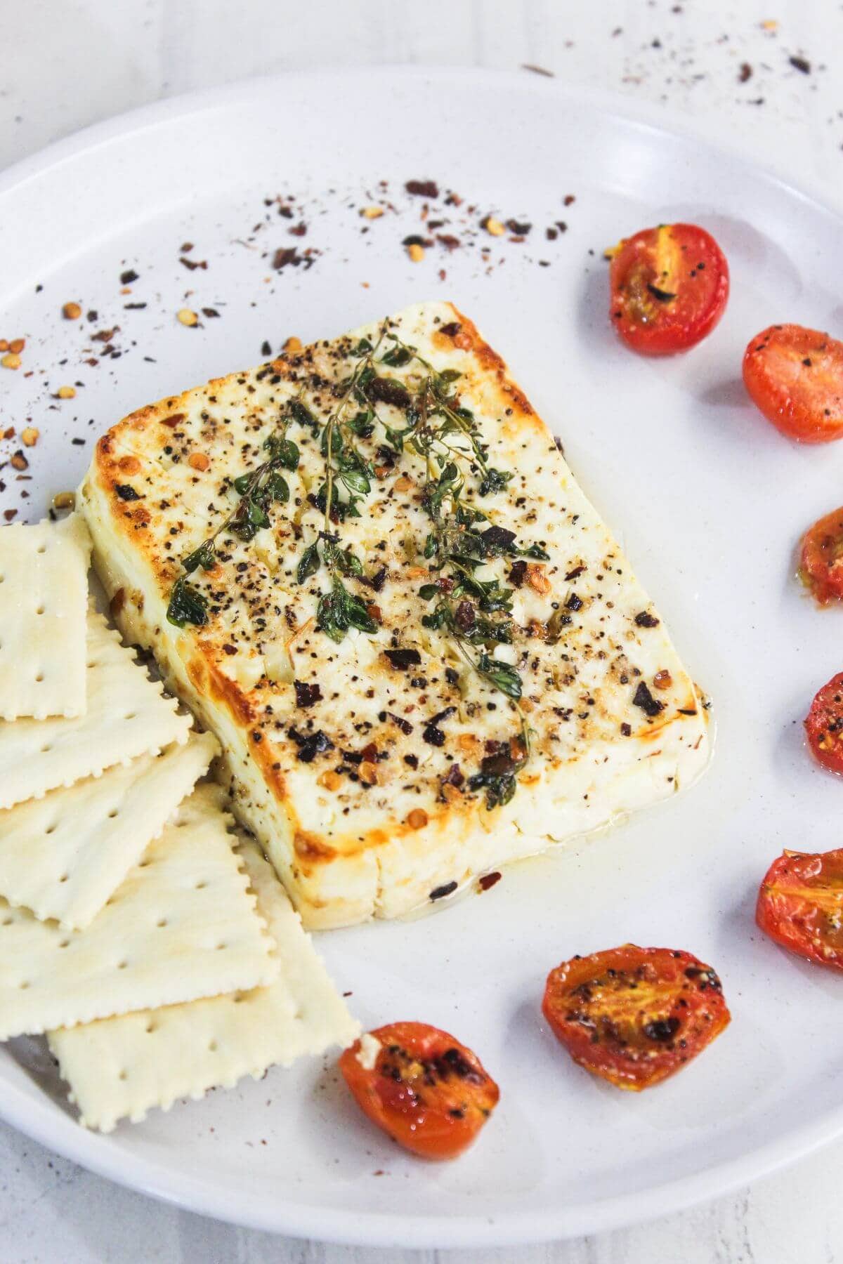 A plate with baked feta cheese topped with herbs and spices, surrounded by crackers and roasted cherry tomato halves.