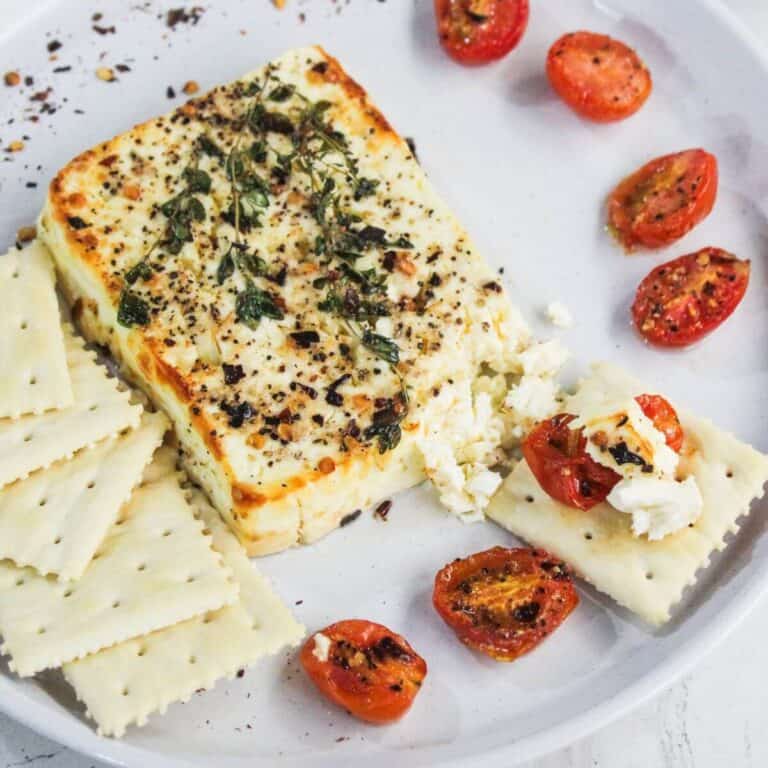 A plate with baked feta cheese topped with herbs, roasted cherry tomatoes, and square crackers, some topped with cheese and tomato pieces.