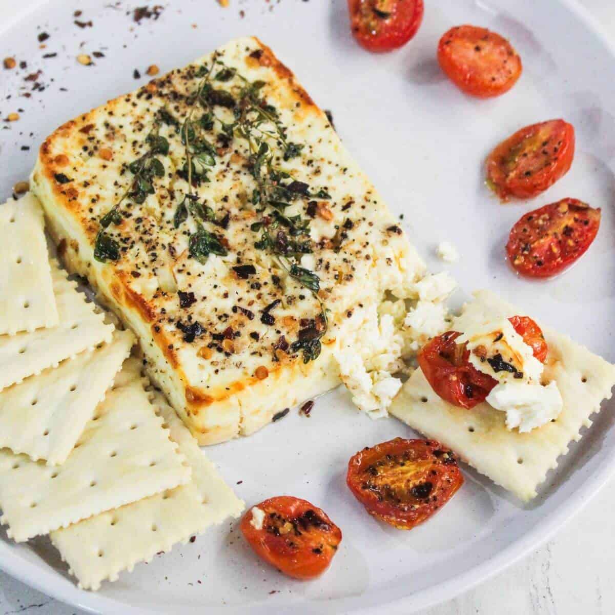 A plate with baked feta cheese topped with herbs, roasted cherry tomatoes, and square crackers, some topped with cheese and tomato pieces.