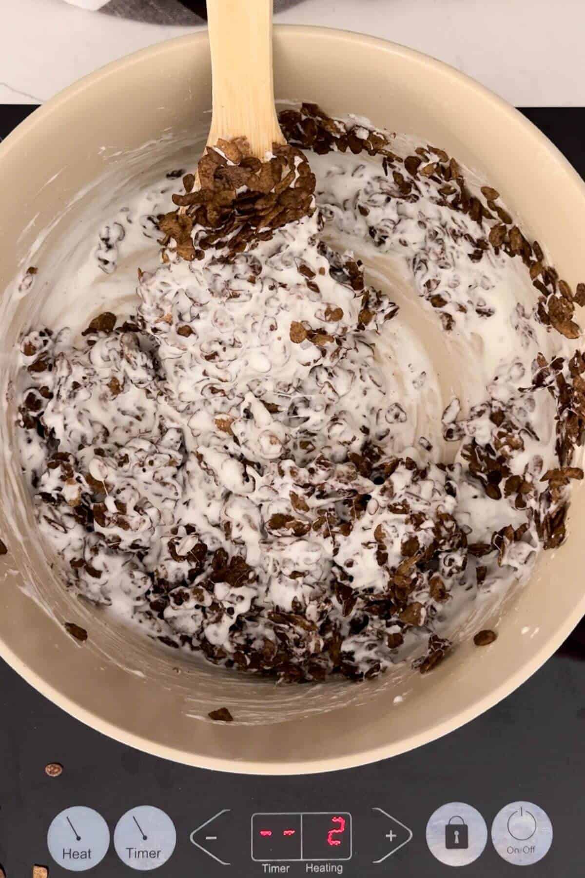 A wooden spoon stirs chocolate cereal flakes coated in melted marshmallow in a beige saucepan on an electric stovetop set to level 2.