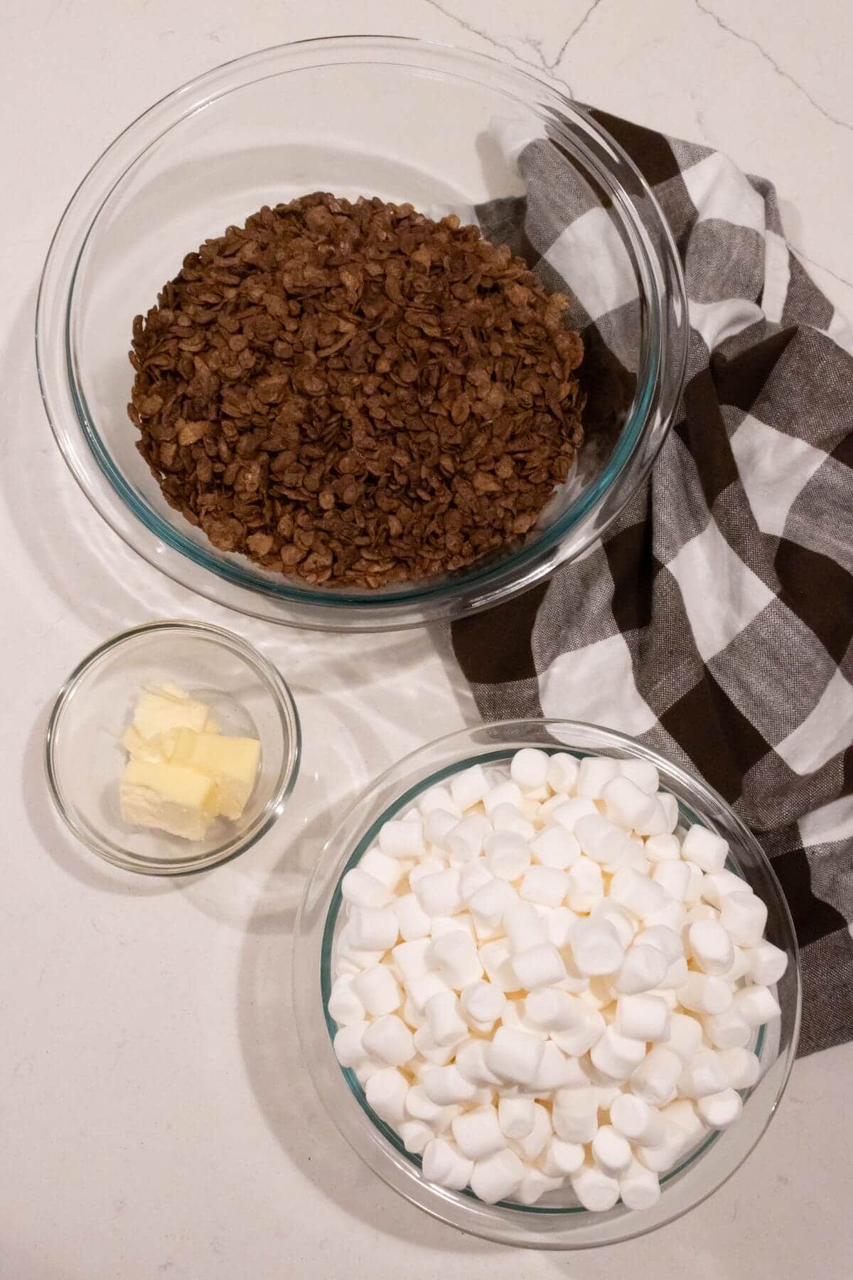 Three glass bowls containing chocolate cereal, butter, and mini marshmallows are placed on a white surface next to a brown and white checkered cloth.