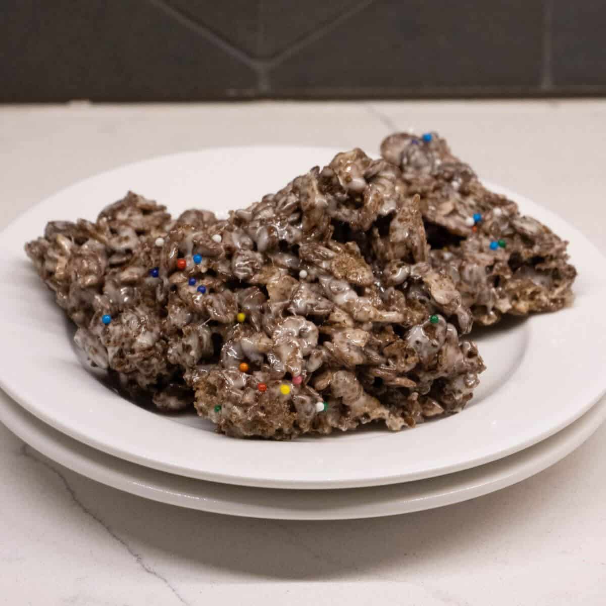 Three chocolate rice cereal treats with colorful sprinkles are stacked on two white plates, set on a marble countertop with a gray tiled backsplash in the background.