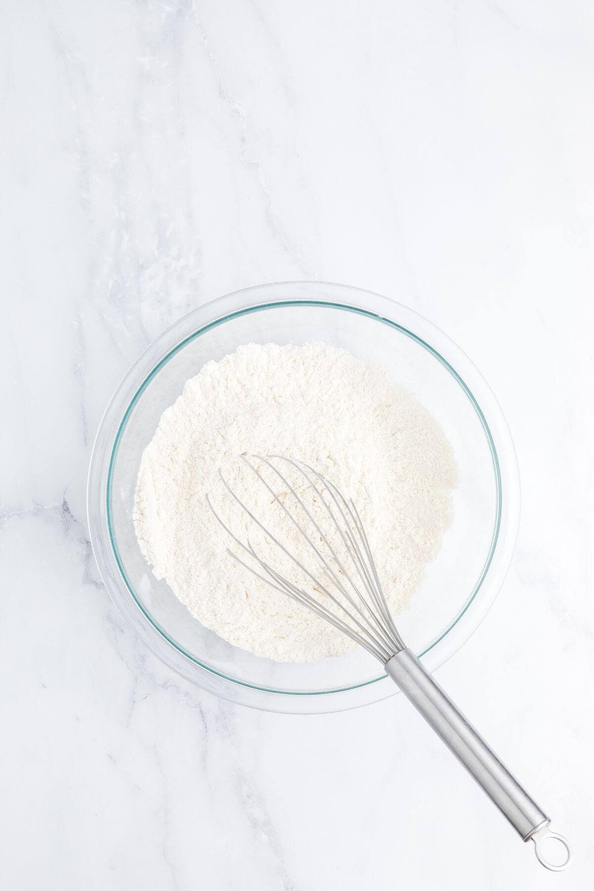A glass bowl filled with flour and a metal whisk resting inside, placed on a white marble surface.