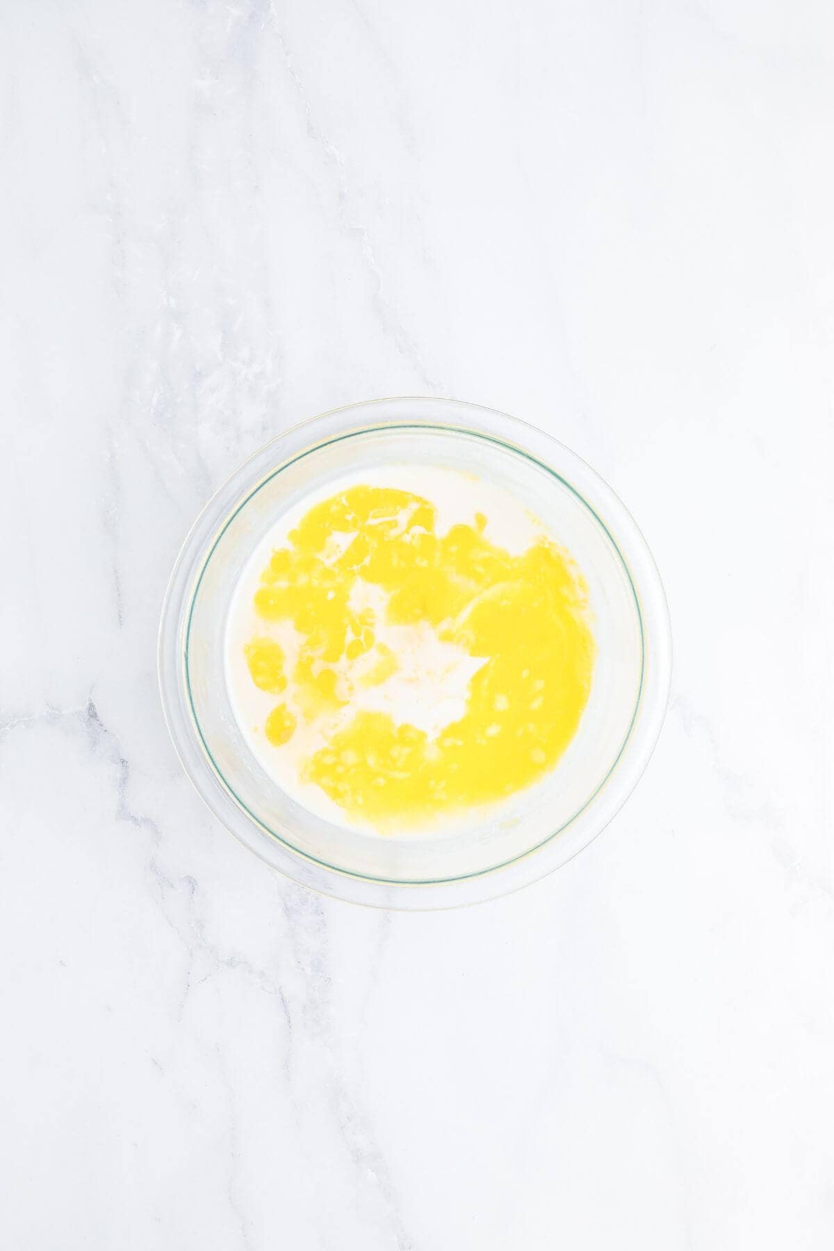 A glass bowl containing a mixture of beaten eggs and milk on a white marble surface.