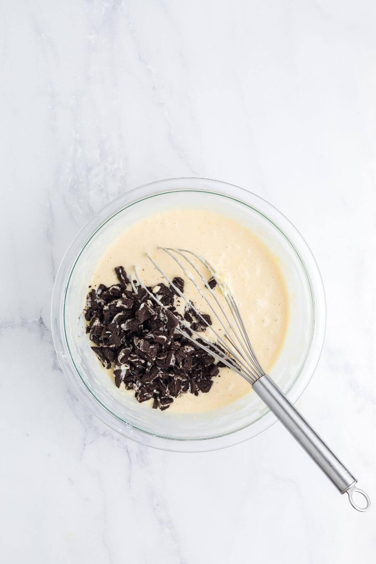 A glass bowl with pale batter and crushed chocolate cookies being mixed with a metal whisk, on a white marble surface.