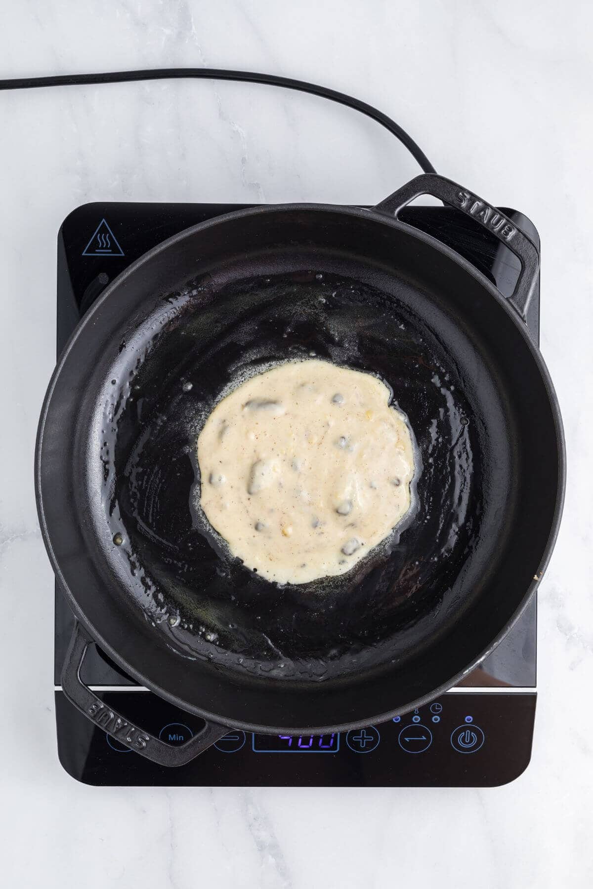 A partially cooked pancake in a black skillet on an electric cooktop with a digital display.