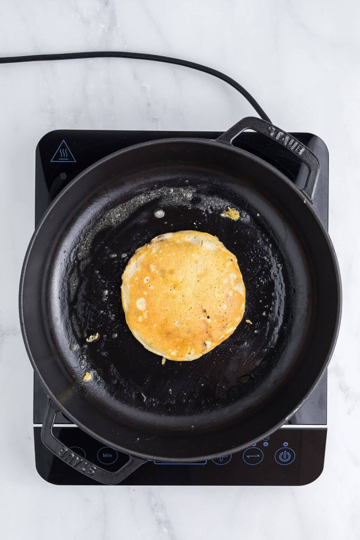 A single pancake cooking in a black skillet on an electric stovetop, viewed from above.