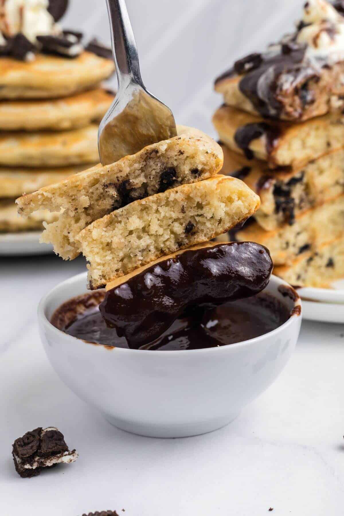 Fork holding a stack of pancakes with chocolate chips, dipping into a bowl of chocolate sauce, with more pancakes topped with cream and sauce in the background.