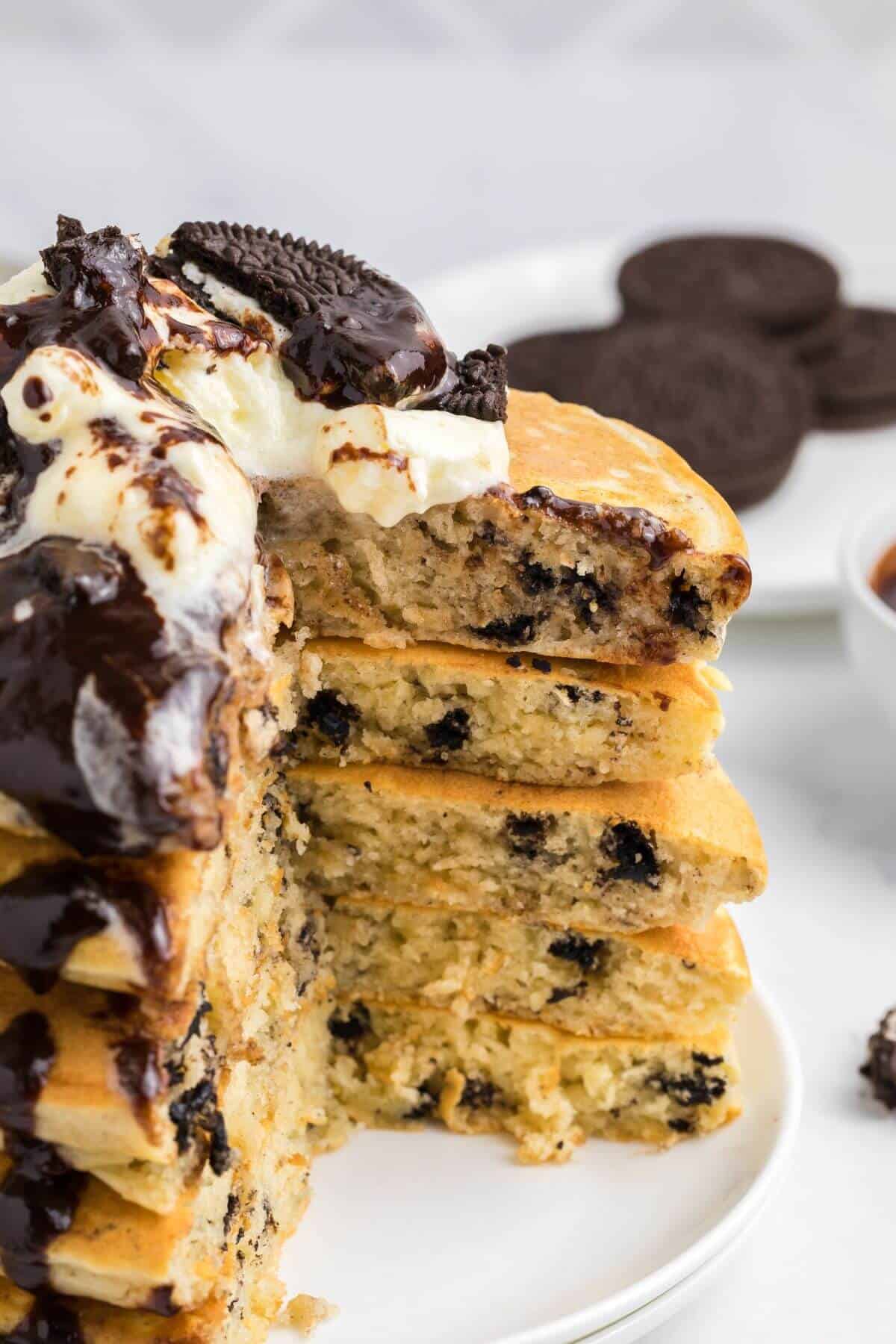 A stack of pancakes with chocolate cookie pieces, topped with whipped cream, chocolate sauce, and more cookies, partially sliced to show the interior.