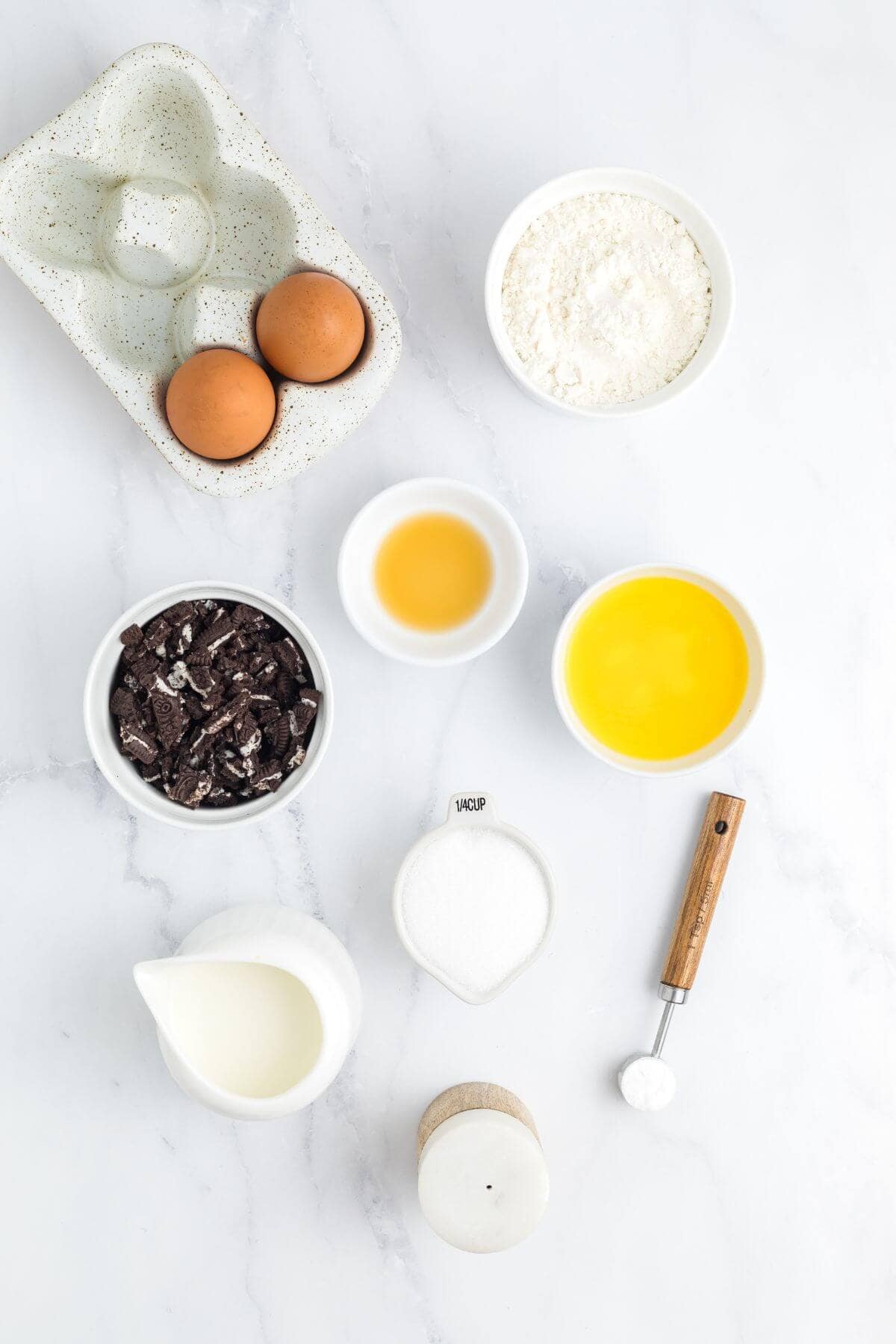 A flat lay of ingredients including eggs, flour, crushed cookies, vanilla, melted butter, milk, sugar, salt, and a measuring spoon on a white surface.