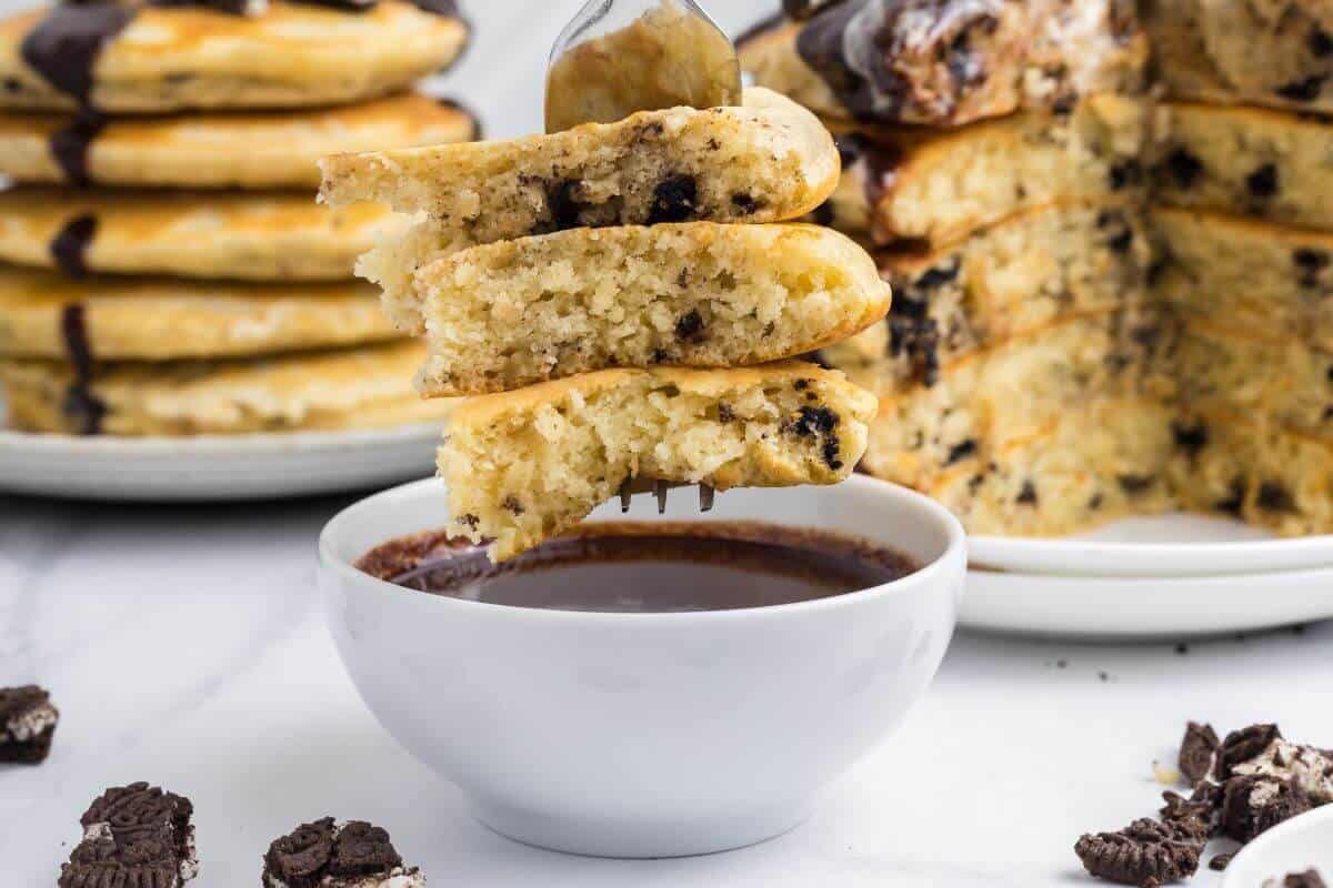 A fork holds three pieces of chocolate chip pancakes above a bowl of chocolate sauce, with more pancakes stacked in the background.