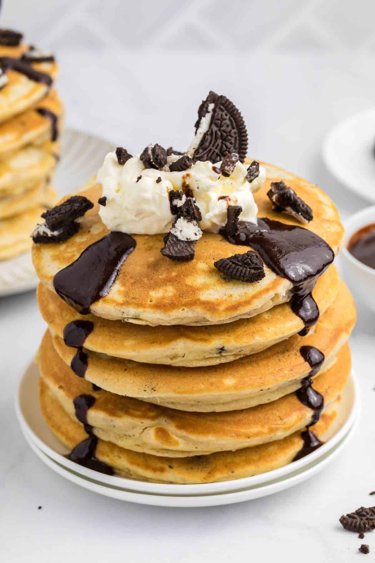 A stack of pancakes topped with whipped cream, chocolate sauce, and crumbled cookies on a white plate.
