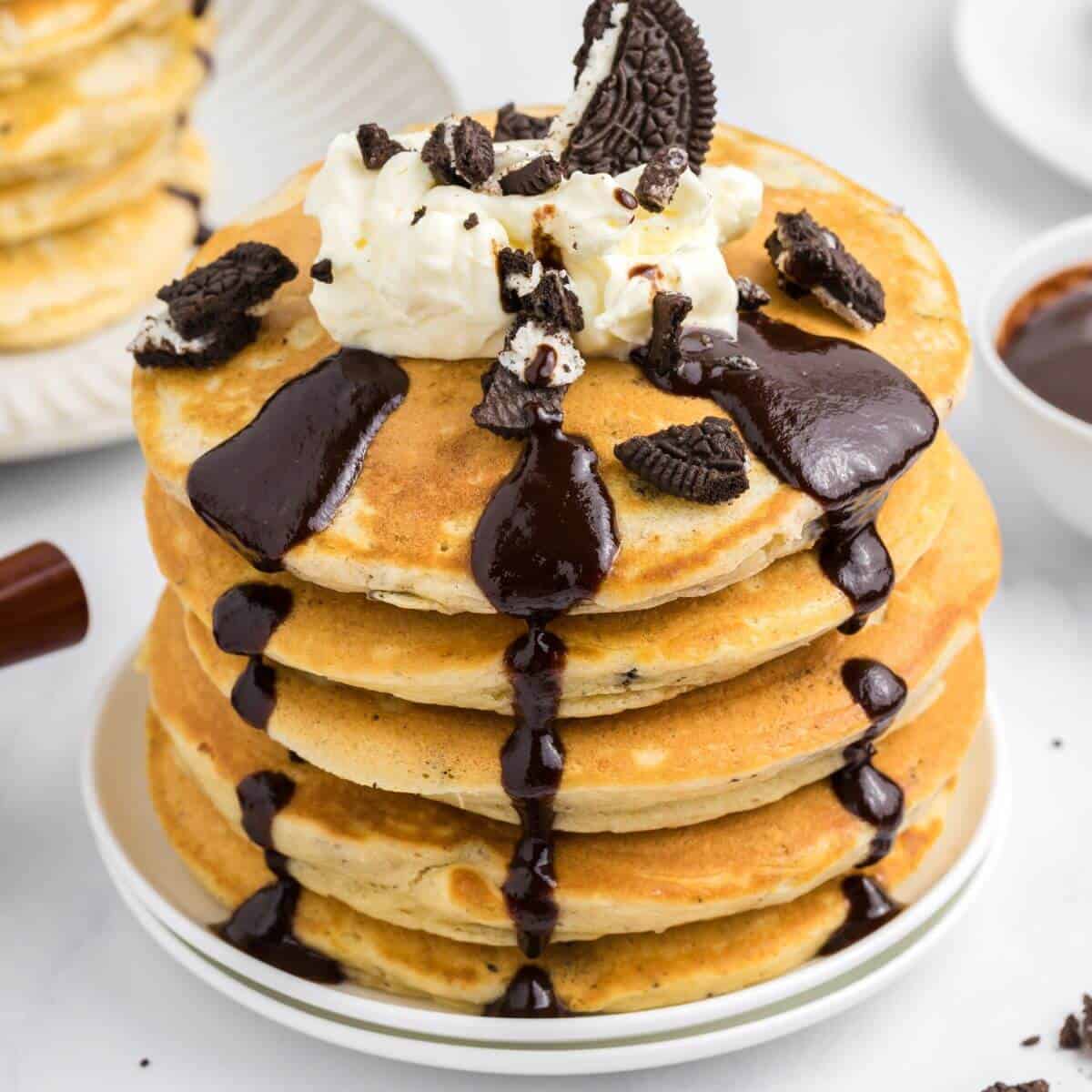 A stack of pancakes topped with whipped cream, chocolate sauce, and crumbled cookies on a white plate.
