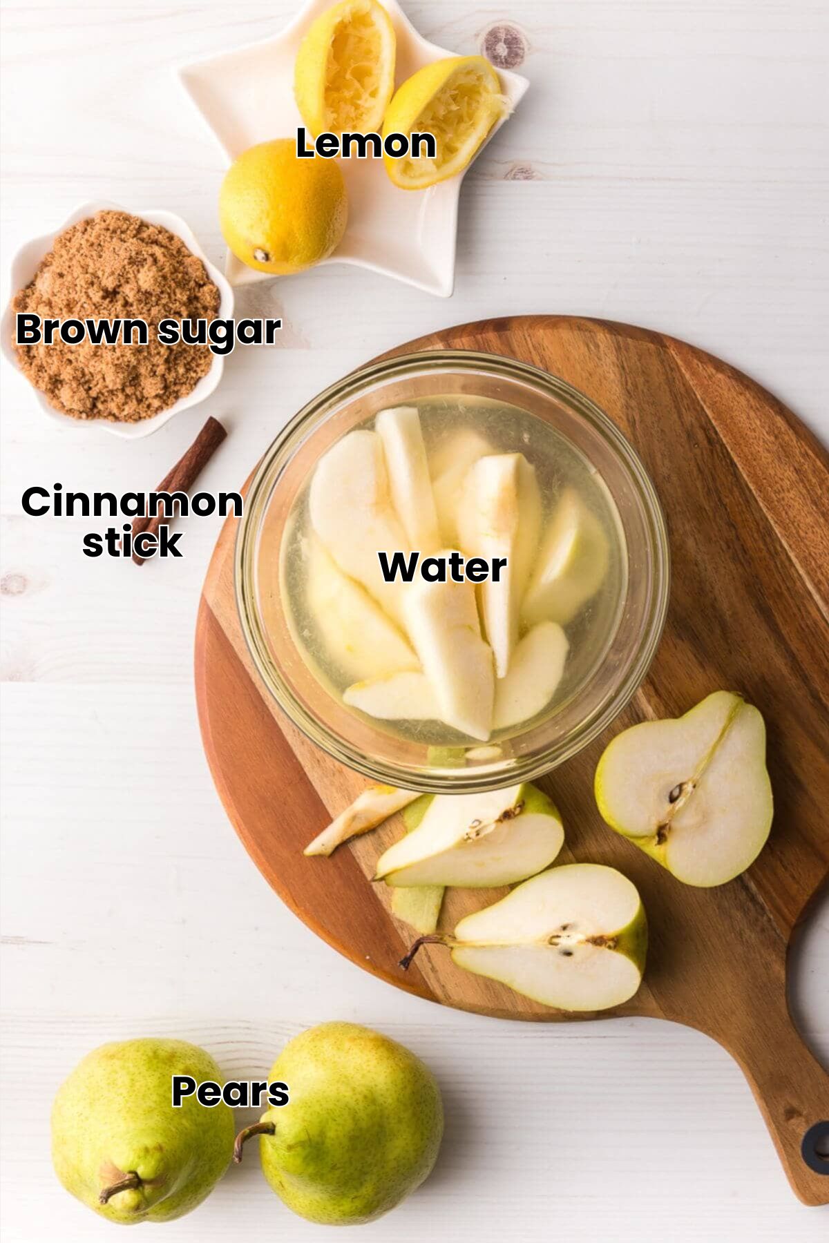 Sliced pears soaking in a bowl, surrounded by whole and halved pears, lemons, brown sugar, and a cinnamon stick on a wooden board and white surface.