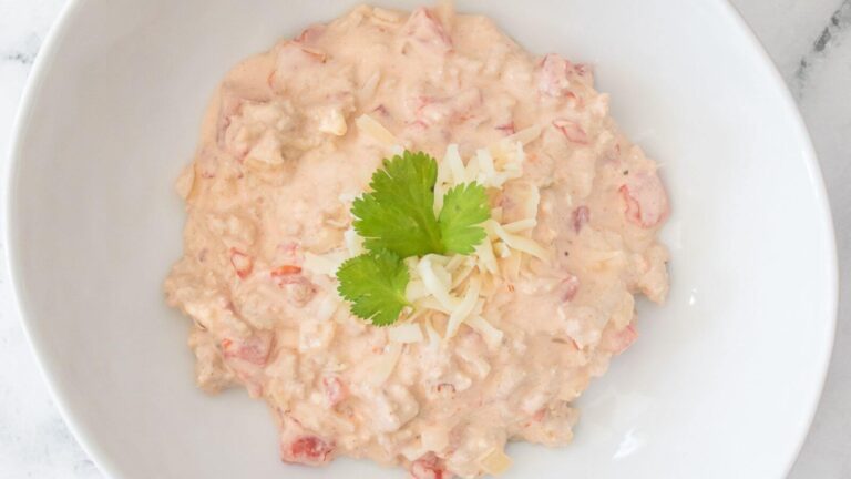 A bowl of creamy dip with diced tomatoes, topped with shredded cheese and a cilantro leaf, served in a white dish.