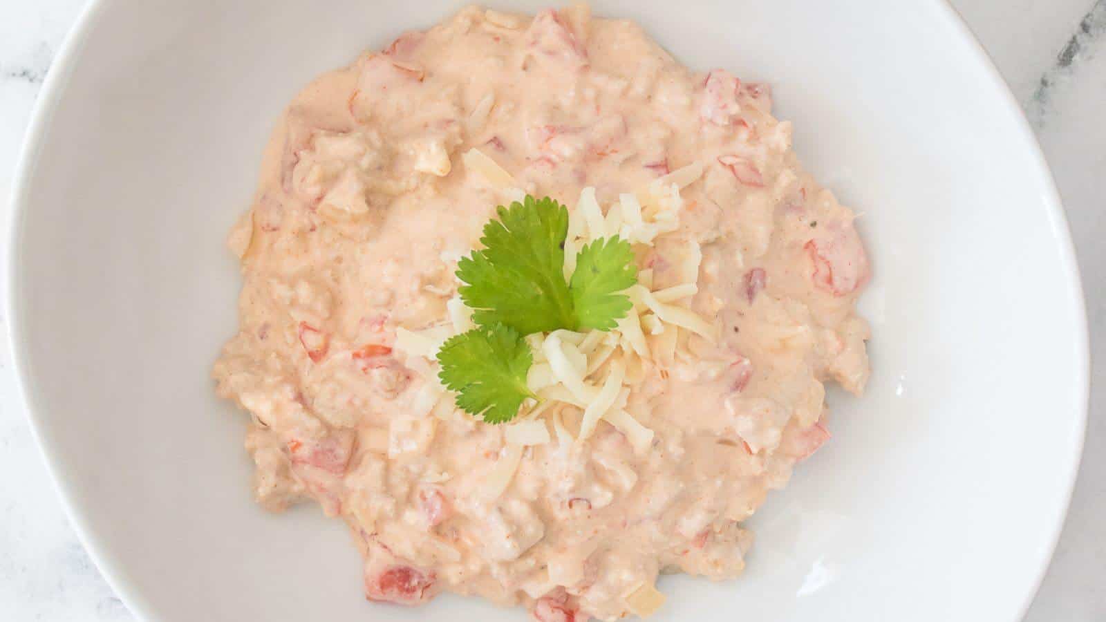 A bowl of creamy dip with diced tomatoes, topped with shredded cheese and a cilantro leaf, served in a white dish.