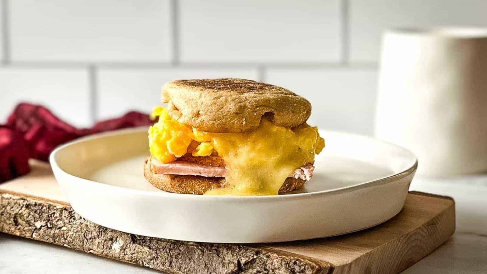 A breakfast sandwich with scrambled eggs, melted cheese, and ham on an English muffin sits on a white plate atop a wooden board.