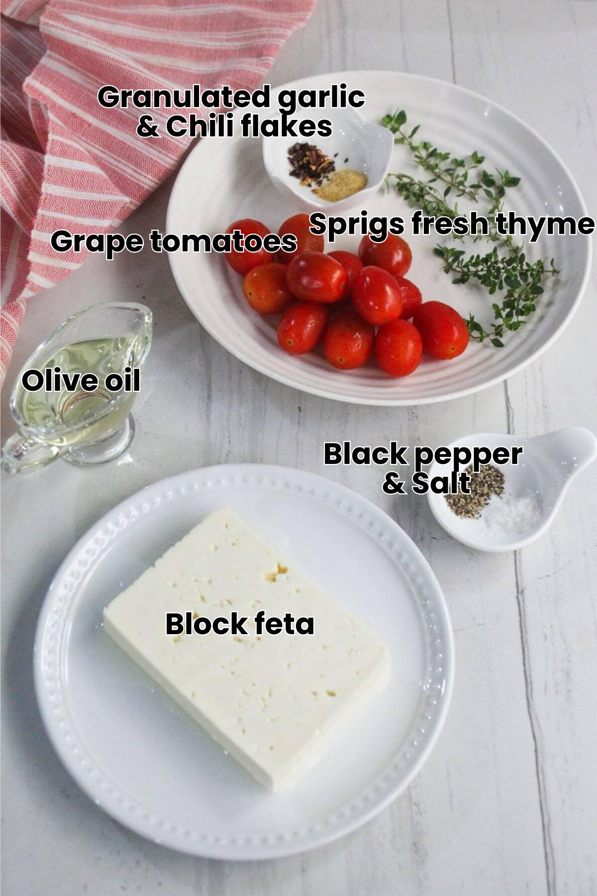 A plate of cherry tomatoes, fresh herbs, and spices, a block of feta cheese on a separate plate, a small glass of olive oil, salt, and pepper on a white surface.