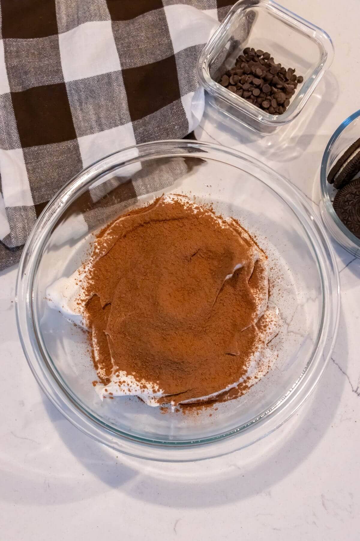 A glass bowl with whipped cream and cocoa powder, next to a small bowl of chocolate chips, cookies, and a brown-and-white checkered cloth.