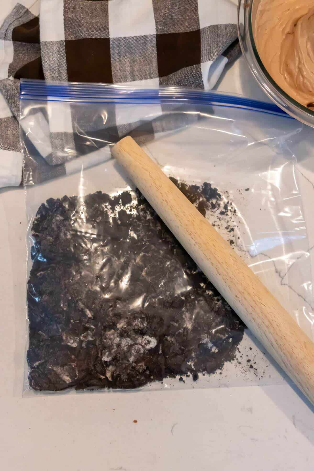 A rolling pin crushing chocolate cookies inside a ziplock bag on a countertop, with a mixing bowl and checkered cloth nearby.