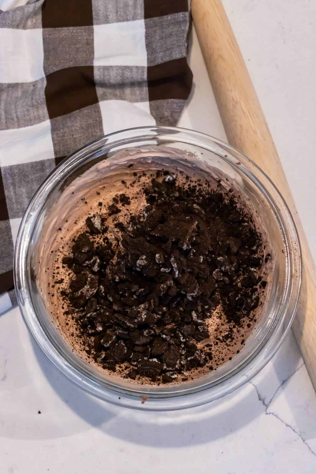A glass bowl filled with chocolate mousse topped with crushed chocolate cookies, next to a checkered cloth and a rolling pin on a white countertop.