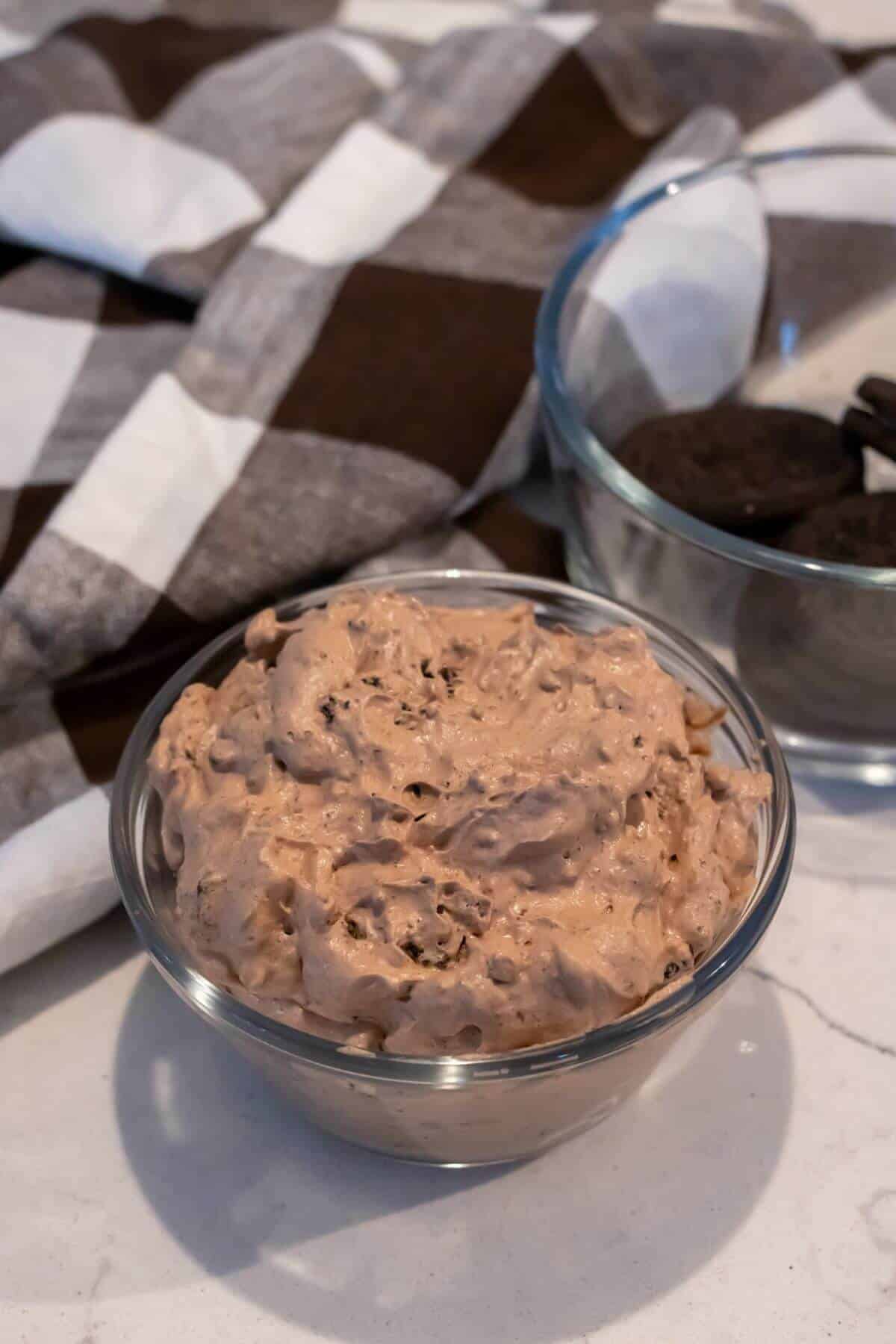 A glass bowl filled with chocolate-flavored creamy dip sits on a countertop next to a bowl of chocolate sandwich cookies and a checkered cloth.