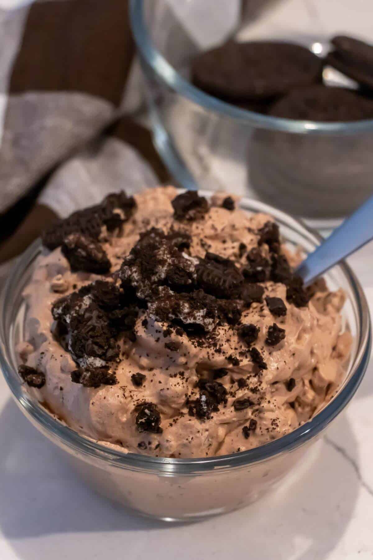 A glass bowl filled with chocolate mousse topped with crushed chocolate cookies, with a spoon inside. Another bowl of whole cookies is in the background.