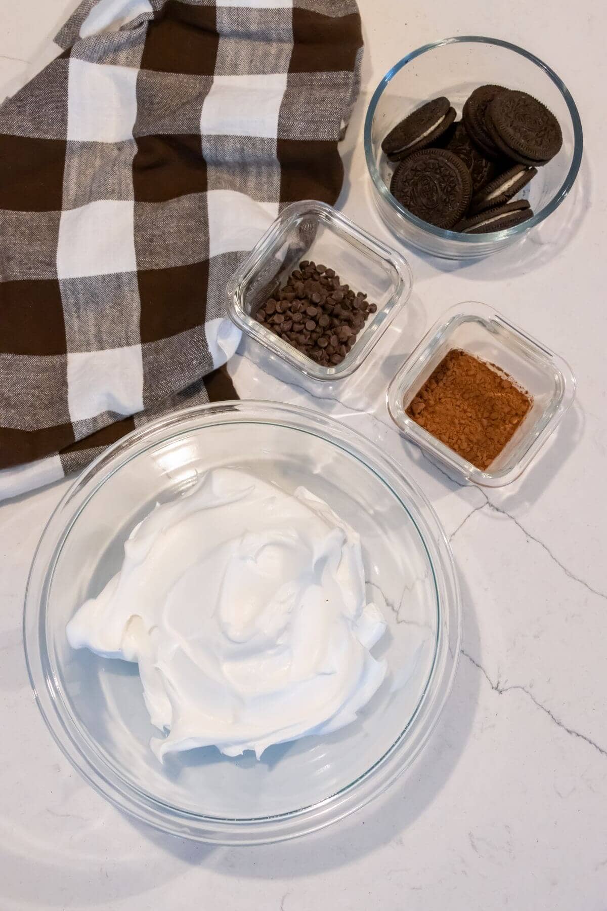 A bowl of whipped cream, a bowl of chocolate sandwich cookies, a bowl of chocolate chips, and a bowl of instant coffee on a counter next to a brown and white checkered cloth.