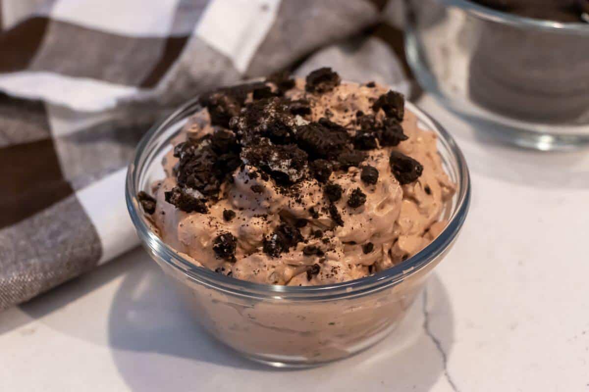 A glass bowl filled with chocolate mousse topped with crushed chocolate cookies, placed on a white surface near a brown and white cloth.